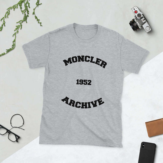 Grey designer t-shirt with "Moncler Archive 1952" text, displayed on a white background with accessories nearby.