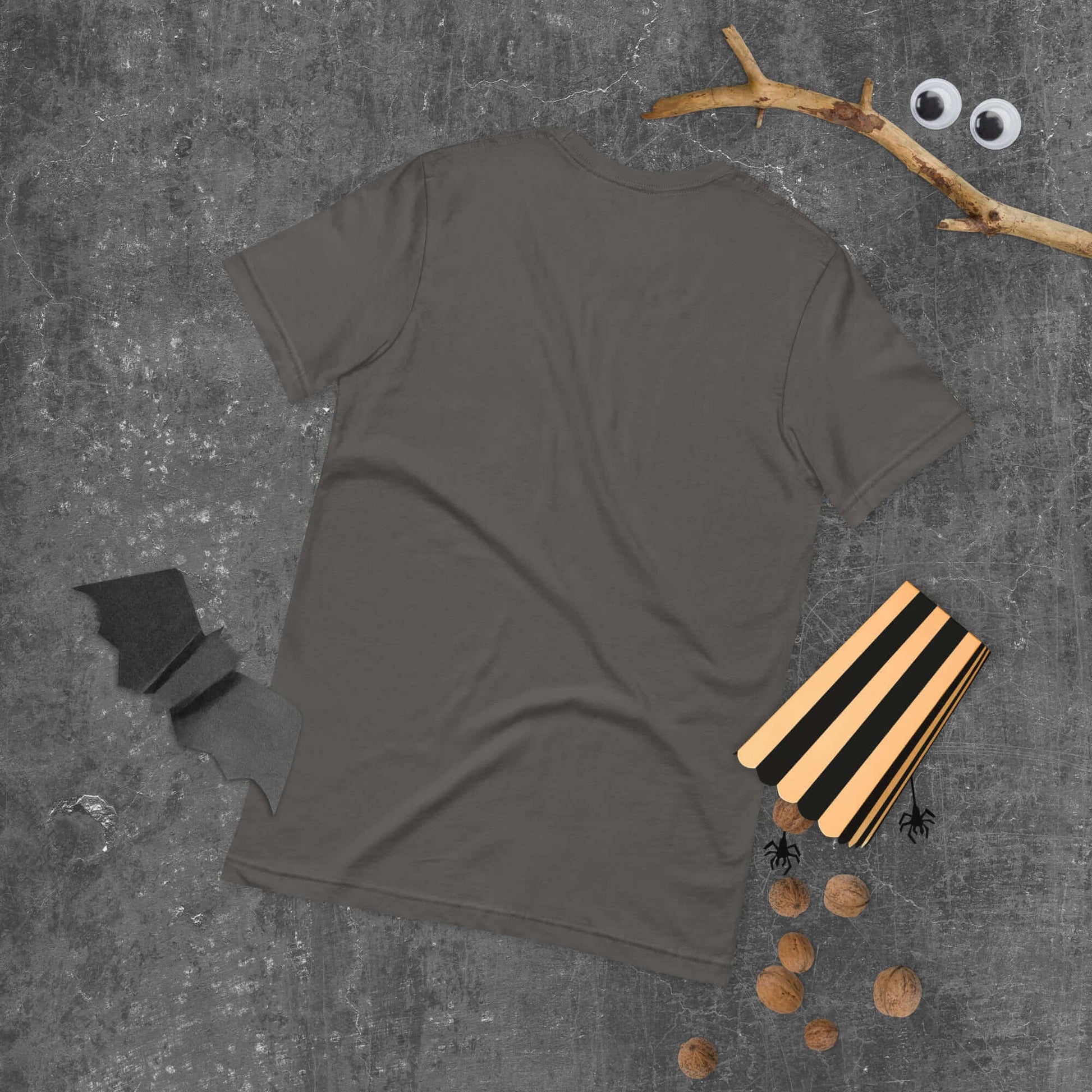 Gray unisex t-shirt with Halloween decorations on concrete background including bat, nuts, small spiders, and twigs with googly eyes.