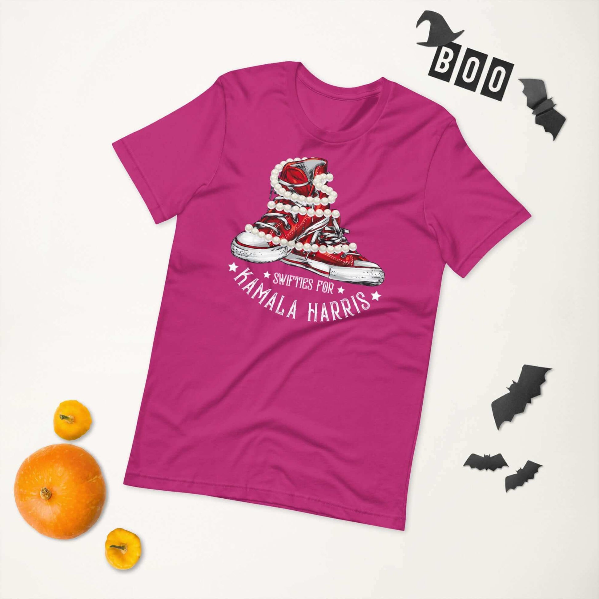 Pink unisex t-shirt with "Swifties for Kamala Harris" design featuring chucks and pearls, surrounded by Halloween decorations.