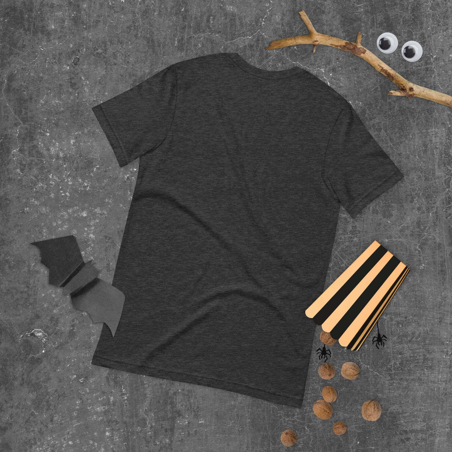 Unisex black t-shirt on a gray surface surrounded by Halloween decorations including a bat cutout and craft materials.