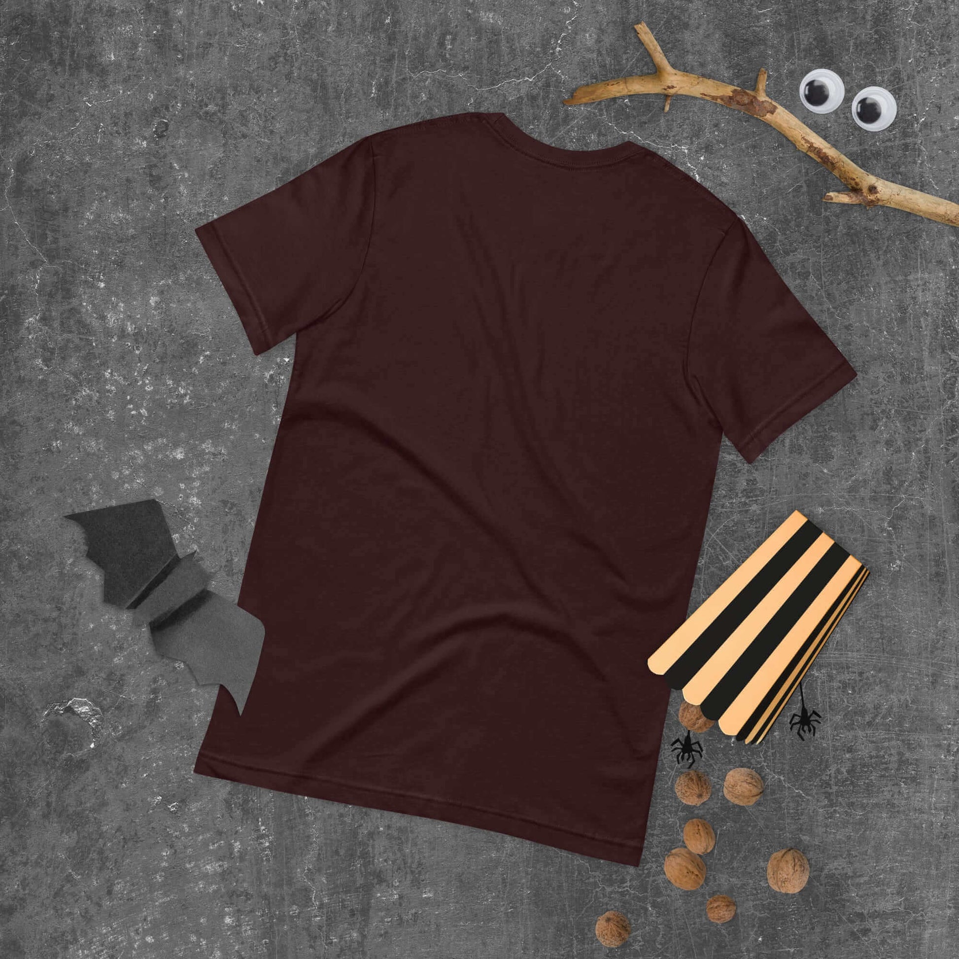 Back view of a brown unisex t-shirt on a textured surface with scattered decorative items.